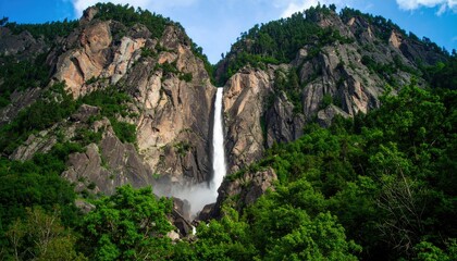 A tall waterfall cascades down a steep, rocky cliff face surrounded by lush green vegetation under a blue sky.