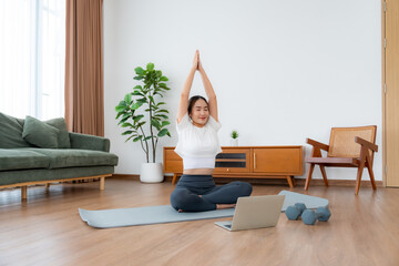 Young Asian woman meditating in lotus position at home, sitting on the floor in fitness clothing....