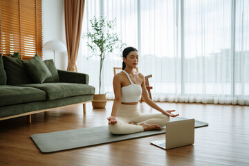 Young Asian woman meditating in lotus position at home, sitting on the floor in fitness clothing....
