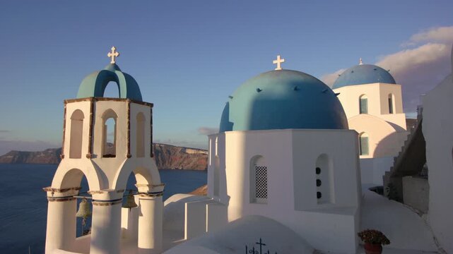 Iconic blue-domed churches overlooking Santorini&rsquo;s dramatic coastline, bathed in warm sunlight with striking Cycladic architecture and Aegean Sea vistas under soft morning skies.