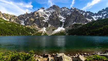 Fototapeta premium A serene lake reflects the towering, snow-capped peaks of a mountain range under a bright blue sky.