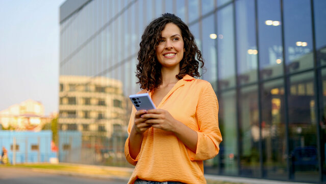 Young businesswoman smiling while using a smartphone, standing confidently in front of a sleek, modern office building in the city