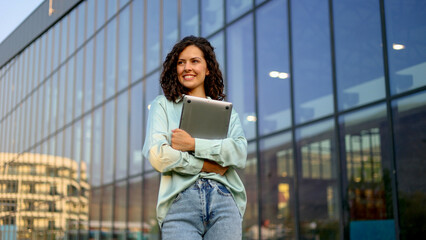 Young businesswoman smiling while holding a laptop, standing confidently in front of a sleek,...