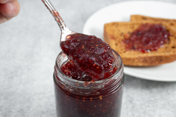 A closeup view of a spoon scooping some raspberry preserves from a jar.