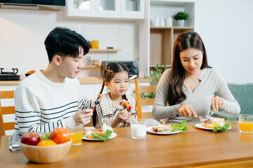 Asian father reads book to daughter at breakfast while mom prepares food. Heartwarming family moment of learning, love, and healthy living in cozy kitchen.