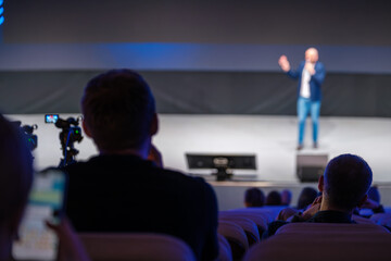Obraz premium Speaker on stage presenting to audience members seen partially, recording equipment placed for capturing moments