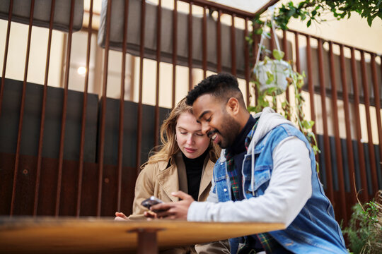 Positive young Arab man using cellphone and showing to girlfriend browsing mobile phone at table on terrace during date. Addicted to devices millennials surfing social media on street
