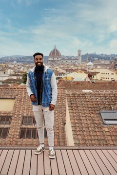 Full body of positive Arab male wearing casual clothes standing on roof of building and leaning on glass fence against old cityscape and looking at camera