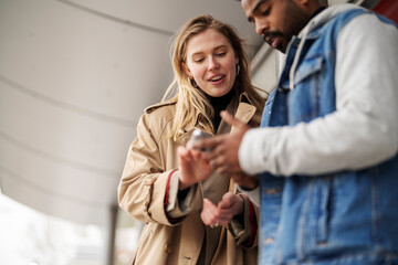 Young woman in trench coat using cellphone and showing to Arab boyfriend while standing together on public transport stop during date