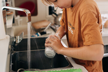 A blond teenage boy washes dishes in the kitchen.