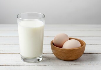 A glass of milk sits beside a bowl of eggs on a white wooden surface