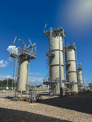 Modern industrial gas processing facility with tall cylindrical structures, pipes, and metallic frameworks under a bright blue sky. An organized site representing energy infrastructure and technology.
