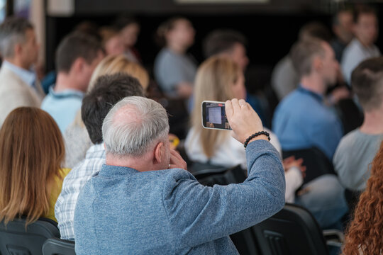 Audience member taking smartphone photo during conference or event with focused view. - Powered by Adobe