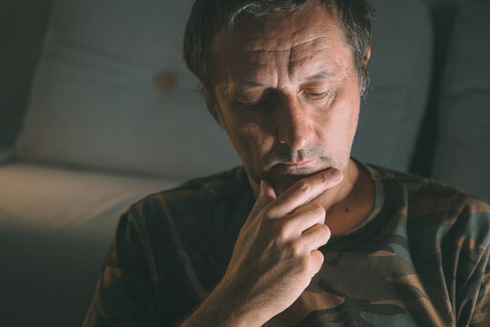 Pensive middle-aged man sitting indoors in low light, deep in thought with hand on chin