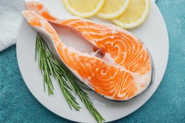 Closeup raw salmon steak with lemon slices and rosemary on white plate over blue background