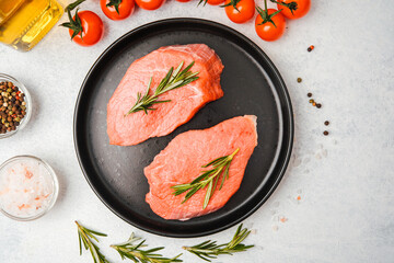 Two raw veal steaks with rosemary on black plate with cooking ingredients around on white surface