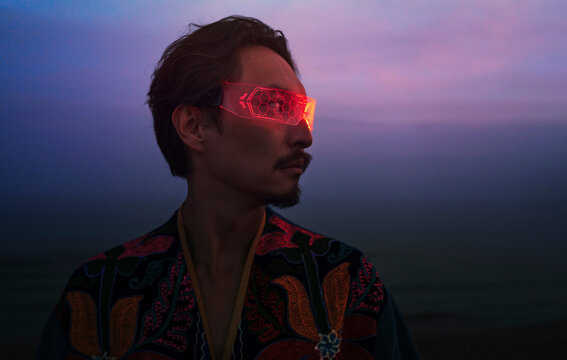 Man in traditional patchwork robe wearing illuminated VR glasses at sunset
