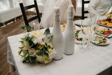 Two bottles of champagne decorated for a wedding celebration stand on a banquet table next to a bouquet of flowers