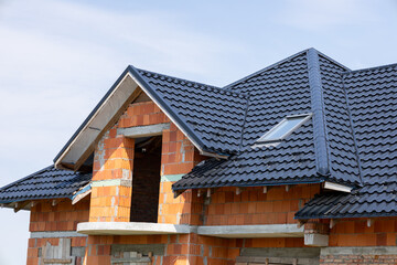 A building under construction showcases a dark roof and brick walls, with clear skies visible above, highlighting progress in a suburban setting