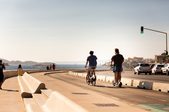 Piste cyclable de la corniche de Marseille en bord de mer avec des personnes sur une trottinette, sur un v&eacute;lo et &agrave; pieds, ville et &icirc;les en arri&egrave;re-plan, Marseille, Sud de la France