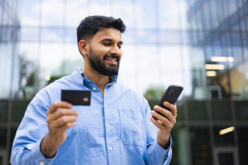 A smiling Indian man holding a credit card and smartphone, likely making an online payment outdoors in front of a modern building.