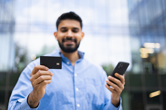 An Indian man smiles holding a credit card and a smartphone in front of a modern building, representing digital banking and financial transactions.