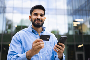 A smiling Indian man is holding a credit card and a smartphone in front of a modern building.