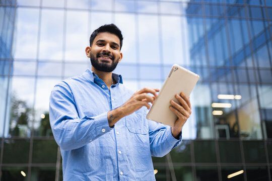 A smiling businessman of Indian descent uses a tablet in front of a modern glass building, conveying a sense of professional success and technological integration.