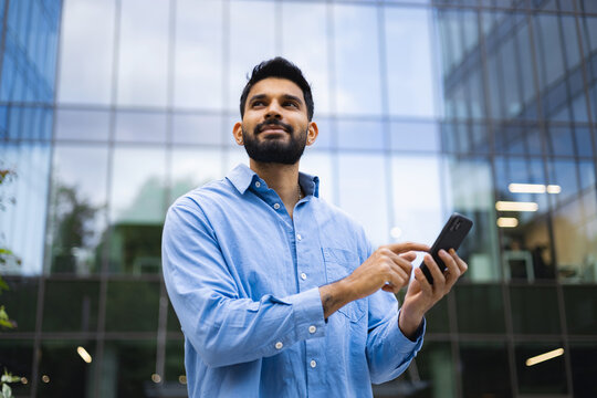 A thoughtful man uses a smartphone outside a modern building.