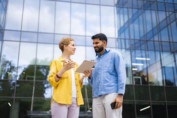 Two business colleagues discuss work using a tablet outside a modern office building, emphasizing collaboration and communication.