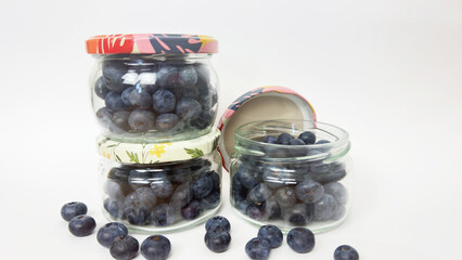 Glass jars filled with fresh blueberries with colorful lids, front view, isolated on white background, healthy organic berries for preservation, natural fruit harvest, vitamin-rich summer food concept