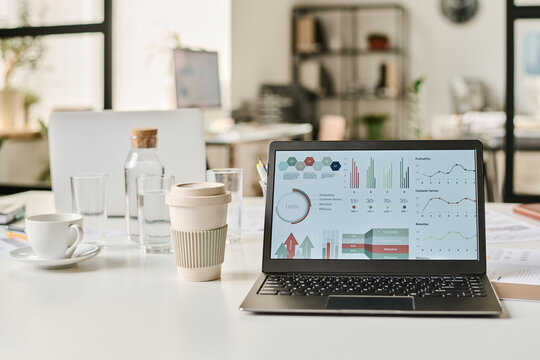 No people shot of laptop with graphs and charts on screen, coffee cup on table in modern office interior, copy space