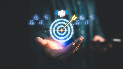Businessman holding digital target with arrow, currency symbols, and handshake icon, representing financial goal achievement, online business, investment success and global business strategy.