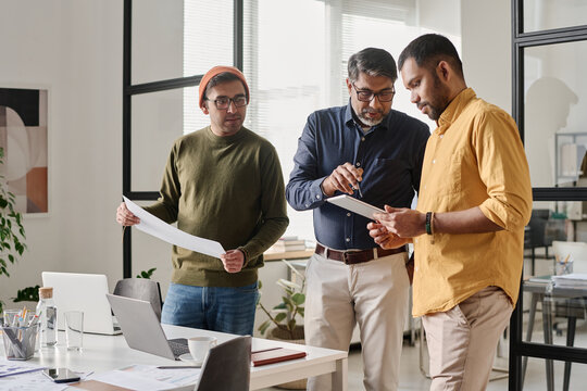 Medium long shot of three South Asian male managers working in team in modern office, using digital tablet and printouts