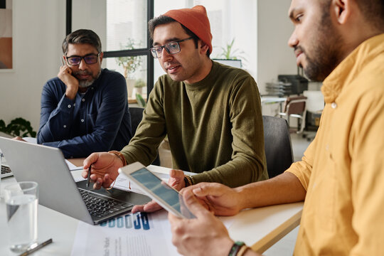 Selective focus shot of three South Asian men sitting at table in office looking at laptop screen and discussing project