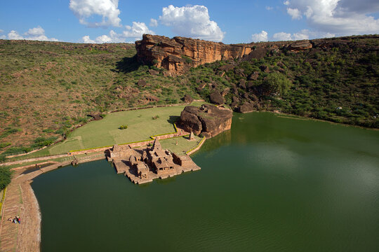 Aerial view of ancient structures and the serene lake nestled against the backdrop of rugged, rust-colored cliffs, Badami, Karnataka, India.