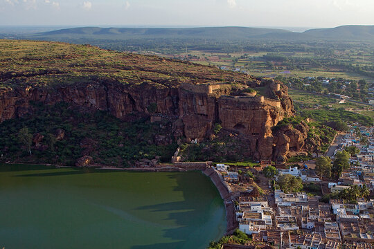 Aerial view of ancient stone fortress walls glowing golden in the afternoon sun, perched atop rugged cliffs overlooking a serene green lake, Badami, Karnataka, India.