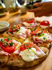 Cooking Pinsa Romana with boiled ham, burrata cheese, tomatoes on wooden kitchen table