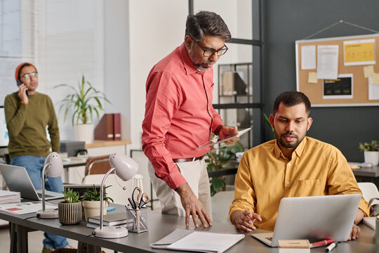 Selective focus shot of Indian men wearing smart casual outfits looking at laptop screen discussing presentation design, their colleague talking on phone in background