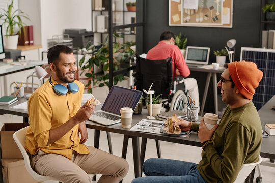 Two joyful Indian male coworkers chatting while having lunch at work in modern office