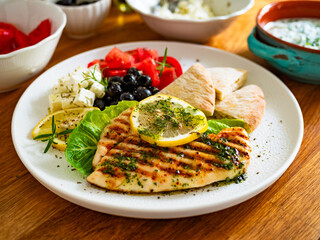 Greek cuisine: grilled chicken breast with lemon, pita bread and greek salad on wooden table	