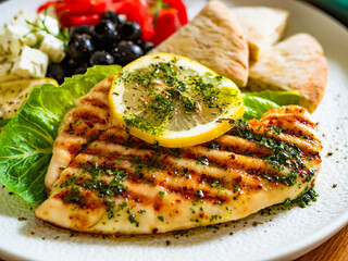 Greek cuisine: grilled chicken breast with lemon, pita bread and greek salad on wooden table	