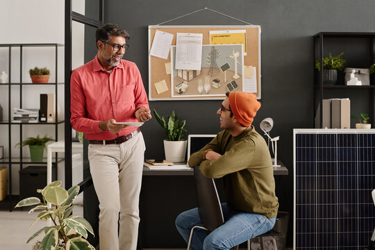 Cheerful mature and young Indian specialists wearing casual clothes standing and sitting in modern office having conversation