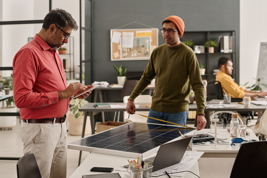 Medium long shot of mature Indian man checking data on digital tablet while his younger colleague measuring size of solar panel at work in office