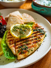 Greek cuisine: grilled chicken breast with lemon, pita bread and greek salad on wooden table	