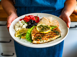 Woman holding plate with grilled chicken breast with lemon, pita bread and greek salad, close-up