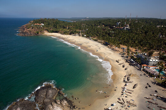 Aerial view of tranquil turquoise waters meet the sandy shores of a tropical beach, fringed by lush green trees and rocky headlands, Kovalam, Kerala, India.