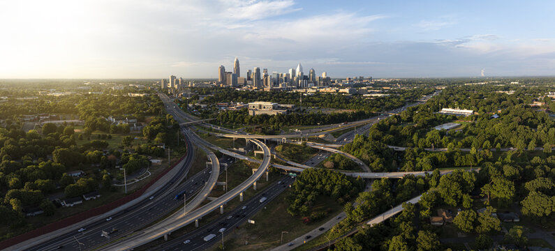 Aerial view of the city skyline contrasts with lush green trees and intricate highway systems, reflecting the warm glow of the setting sun, Charlotte, North Carolina, United States.