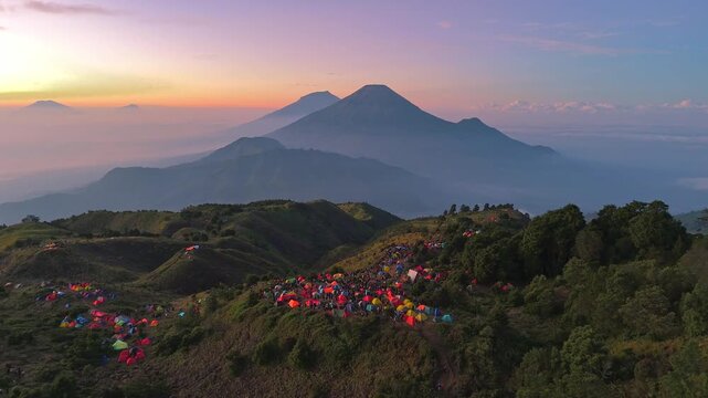 Aerial view of colorful tents scattered across a mountain campsite during sunrise, with majestic volcanic peaks in the background and a calm morning atmosphere. Mount prau landscape, Indonesia.