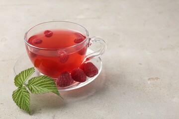 Tasty raspberry tea in glass cup, berries and green leaves on light grey table, closeup. Space for text